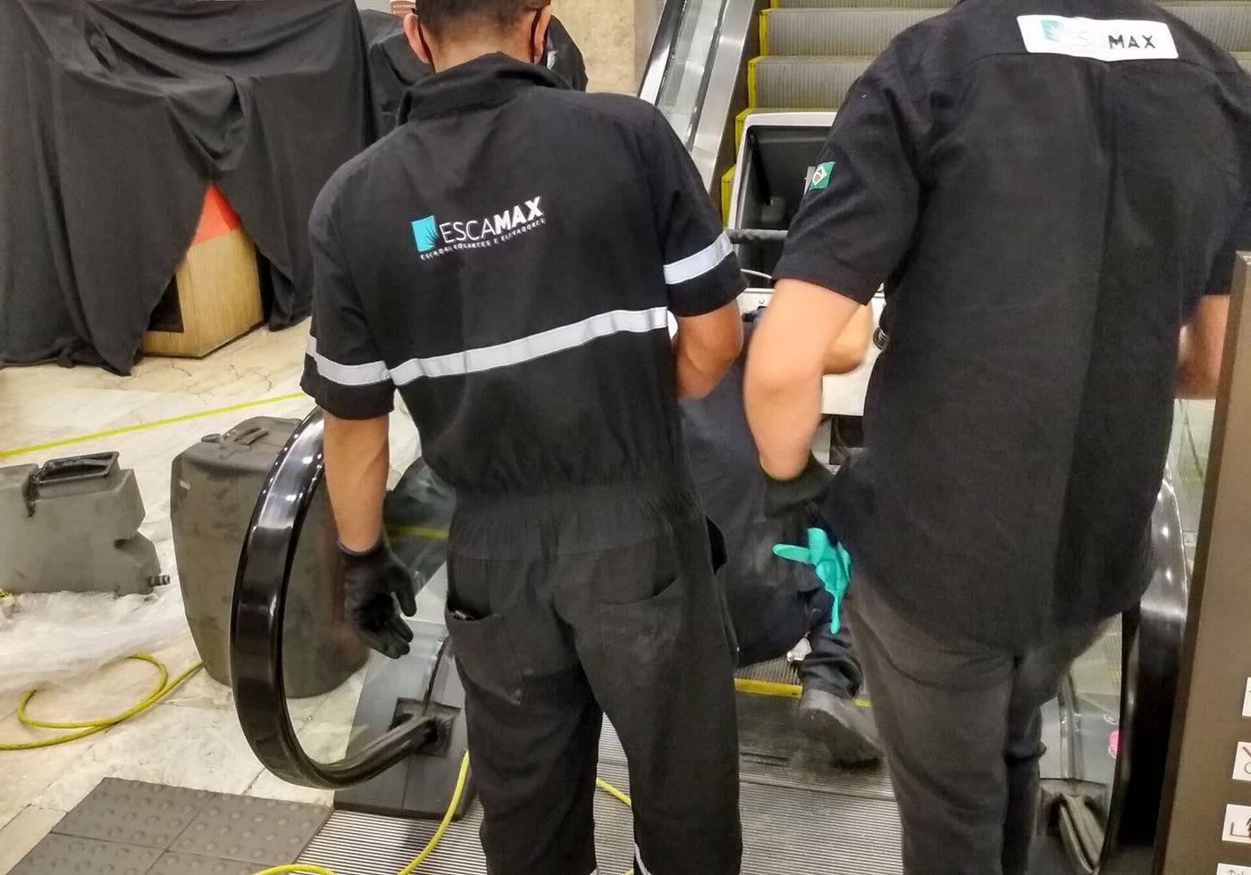 Dois técnicos da Escamax de uniforme trocando o corrimão de uma escada rolante em operação.